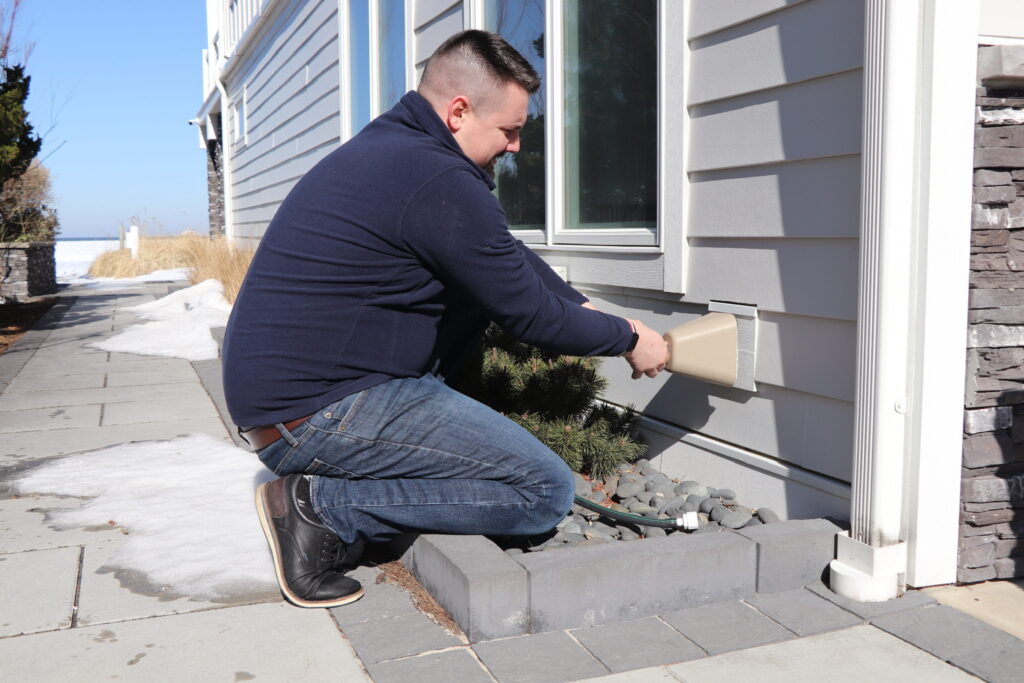 A man kneels outside a house, reaching into a vent on the exterior wall near a window, with snow on the ground and landscaping stones nearby.