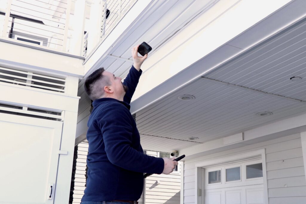A person in a navy jacket stands outside a house, reaching up to install or adjust a security camera under the roof eaves. The house exterior is white with a garage door visible in the background.