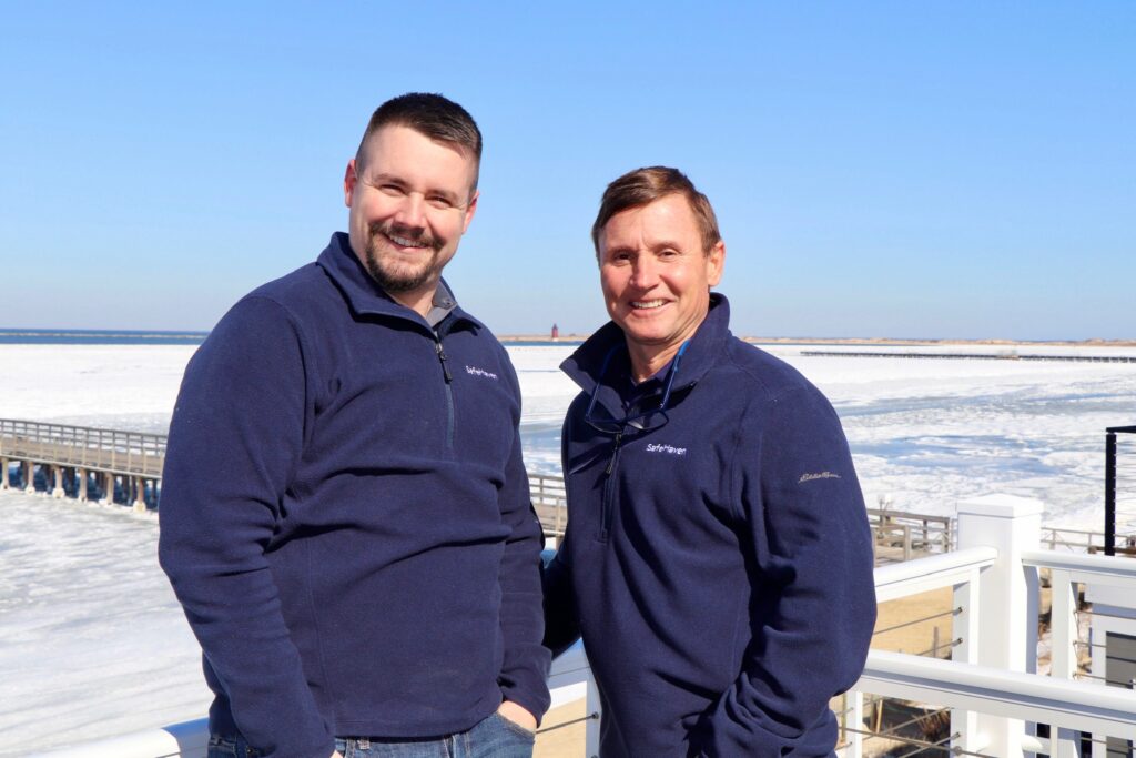 Two men in navy blue sweaters smile while standing on a snowy pier beside a frozen body of water under a clear blue sky